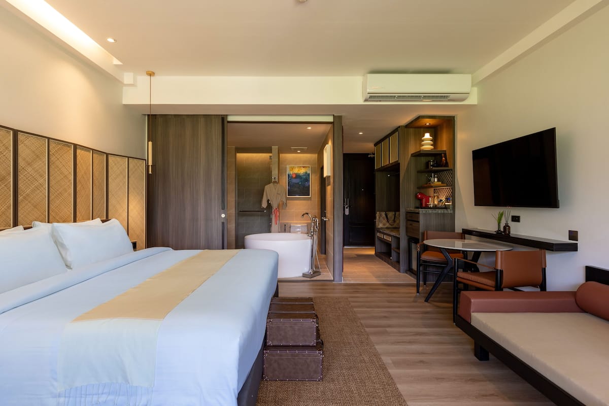 A hotel room with a large bed, a sofa, a dining table, a TV, a bathroom with a bathtub, and a kitchen area.
