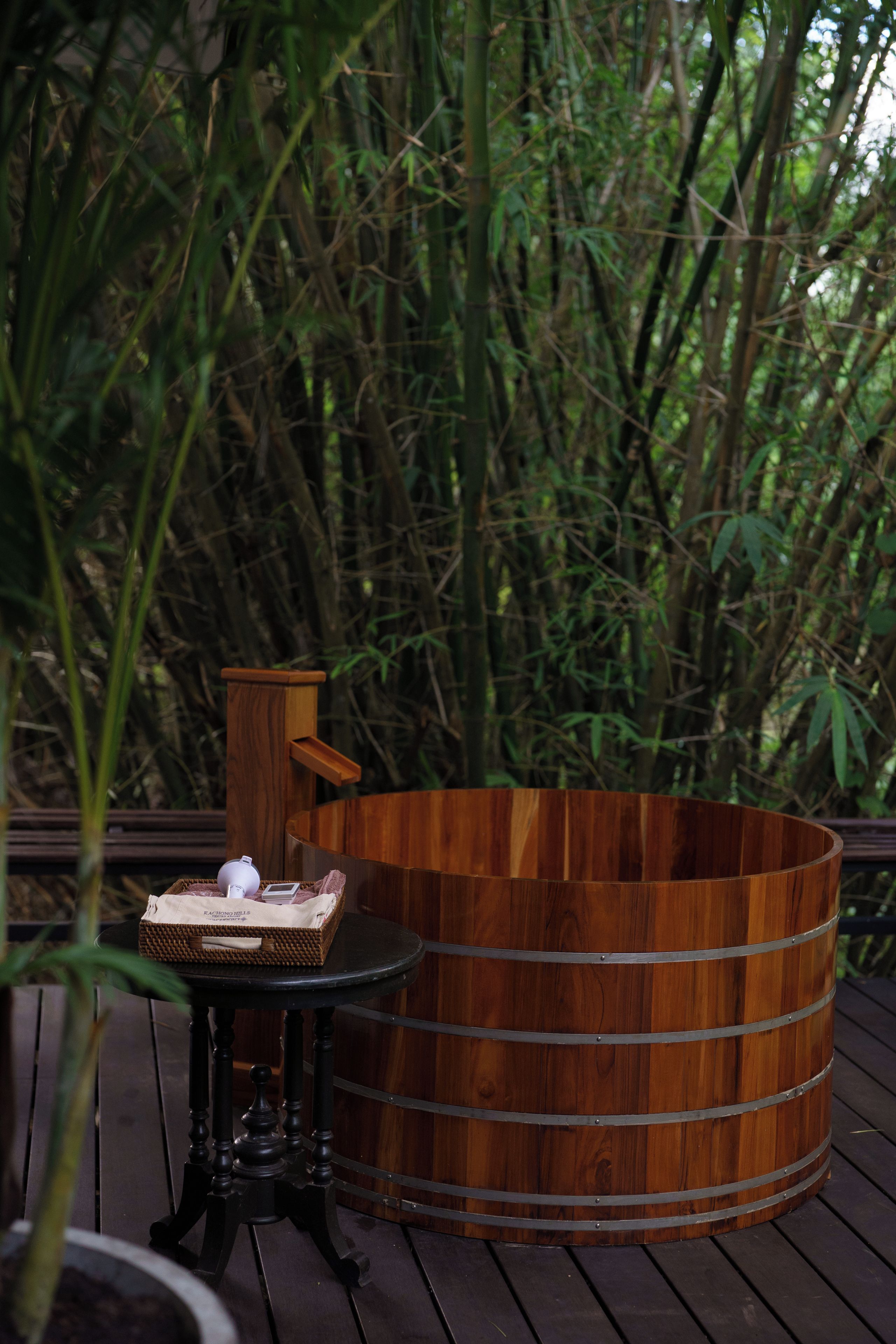A wooden hot tub on a deck with a table and a basket nearby.