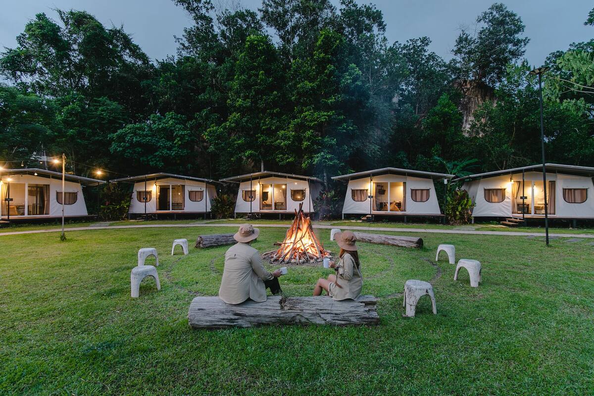 A campsite with a central fire, surrounded by white tents with windows, set in a grassy area with trees in the background.