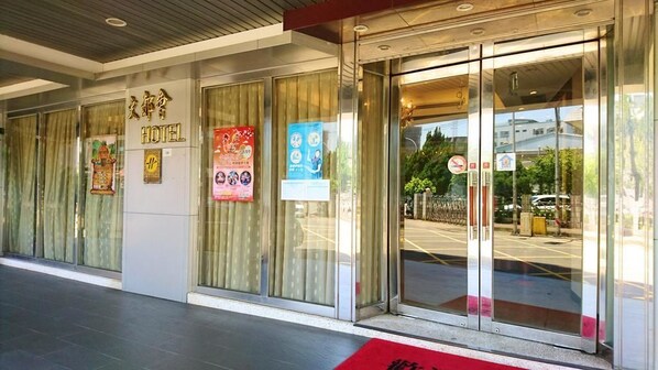 A hotel entrance with glass doors, a red carpet, and various informational posters on the wall.