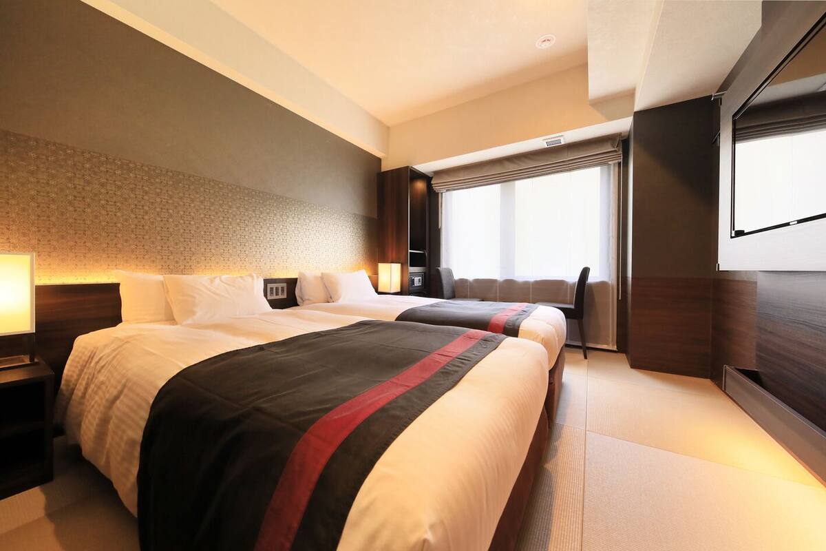 A hotel room with two beds, a large window with curtains, a flat-screen TV, and a wall-mounted air conditioning unit.