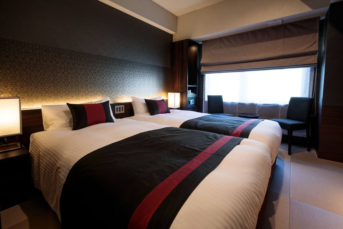 A hotel room with two beds, a large window with curtains, a nightstand, and a chair.