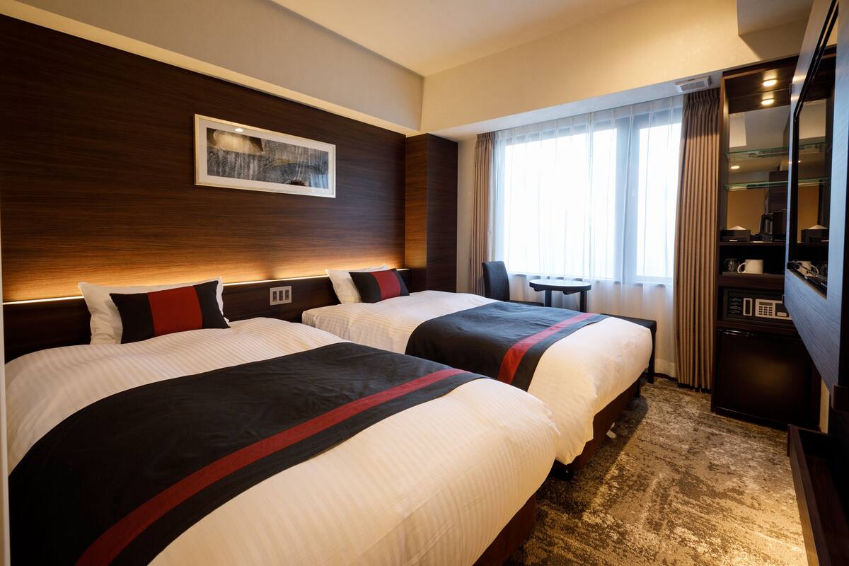 A hotel room with two beds, a desk, a chair, a microwave, and a large window with curtains.
