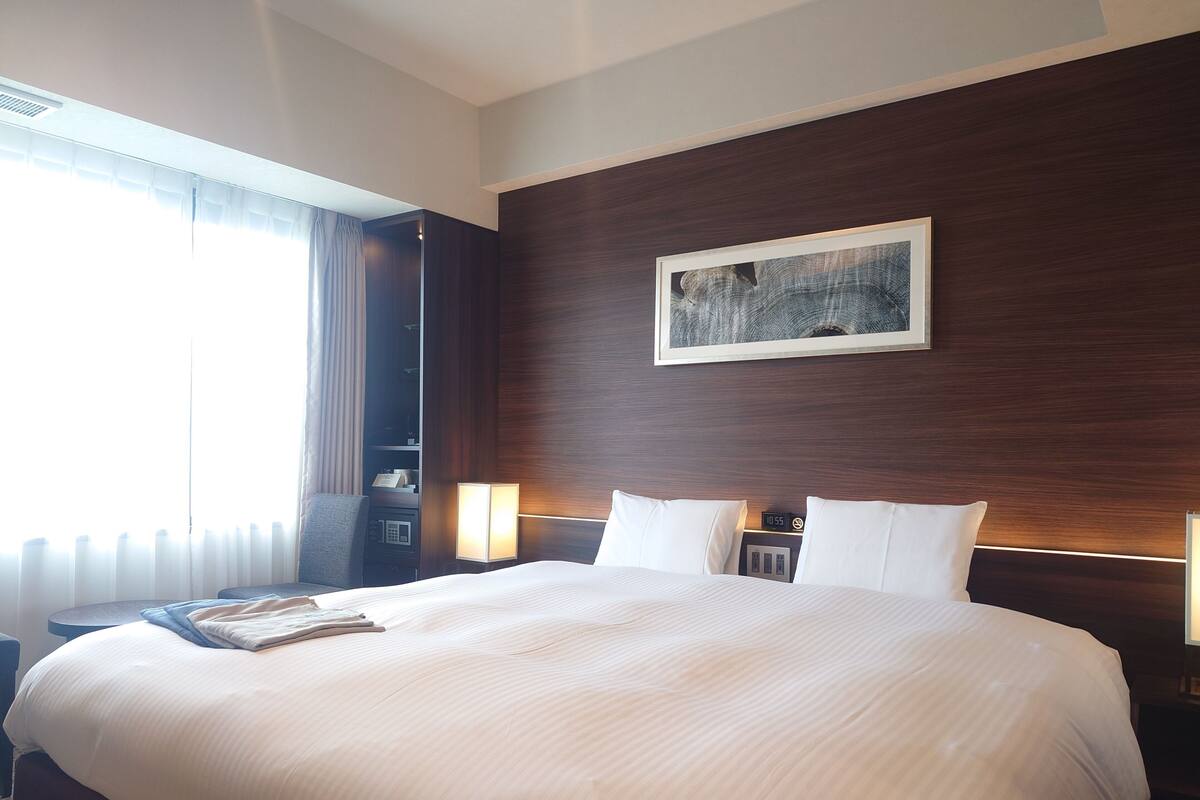A large bed with a headboard, bedside lamps, a clock, and a painting on the wall.