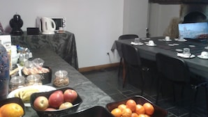 Un coin repas avec une longue table dressée pour un repas, divers fruits disposés sur la table, et une cheminée en arrière-plan.