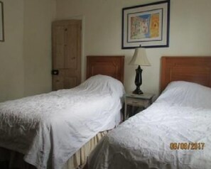 A hotel room with two single beds, a wooden door, a small table with a lamp, and a framed picture on the wall.
