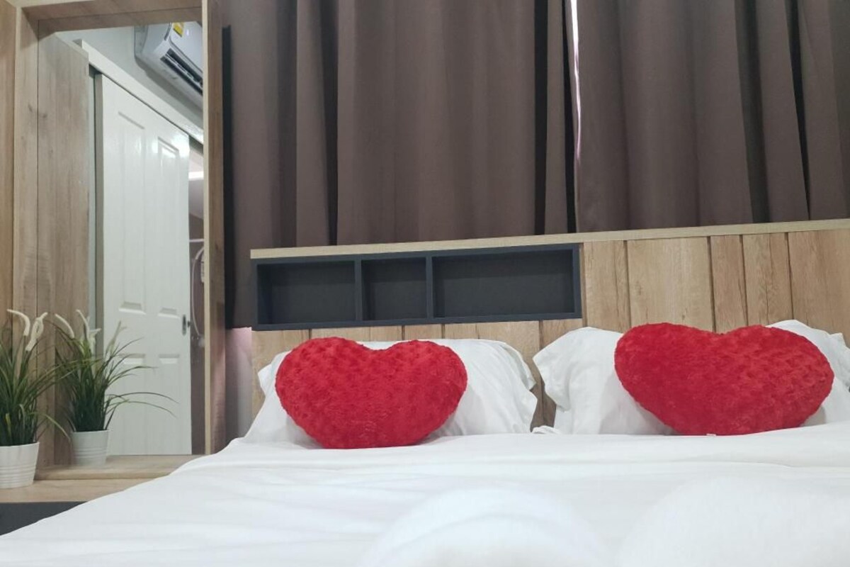 Una cama con ropa blanca y dos almohadas rojas con forma de corazón, un cabecero de madera y una planta en maceta al lado.
