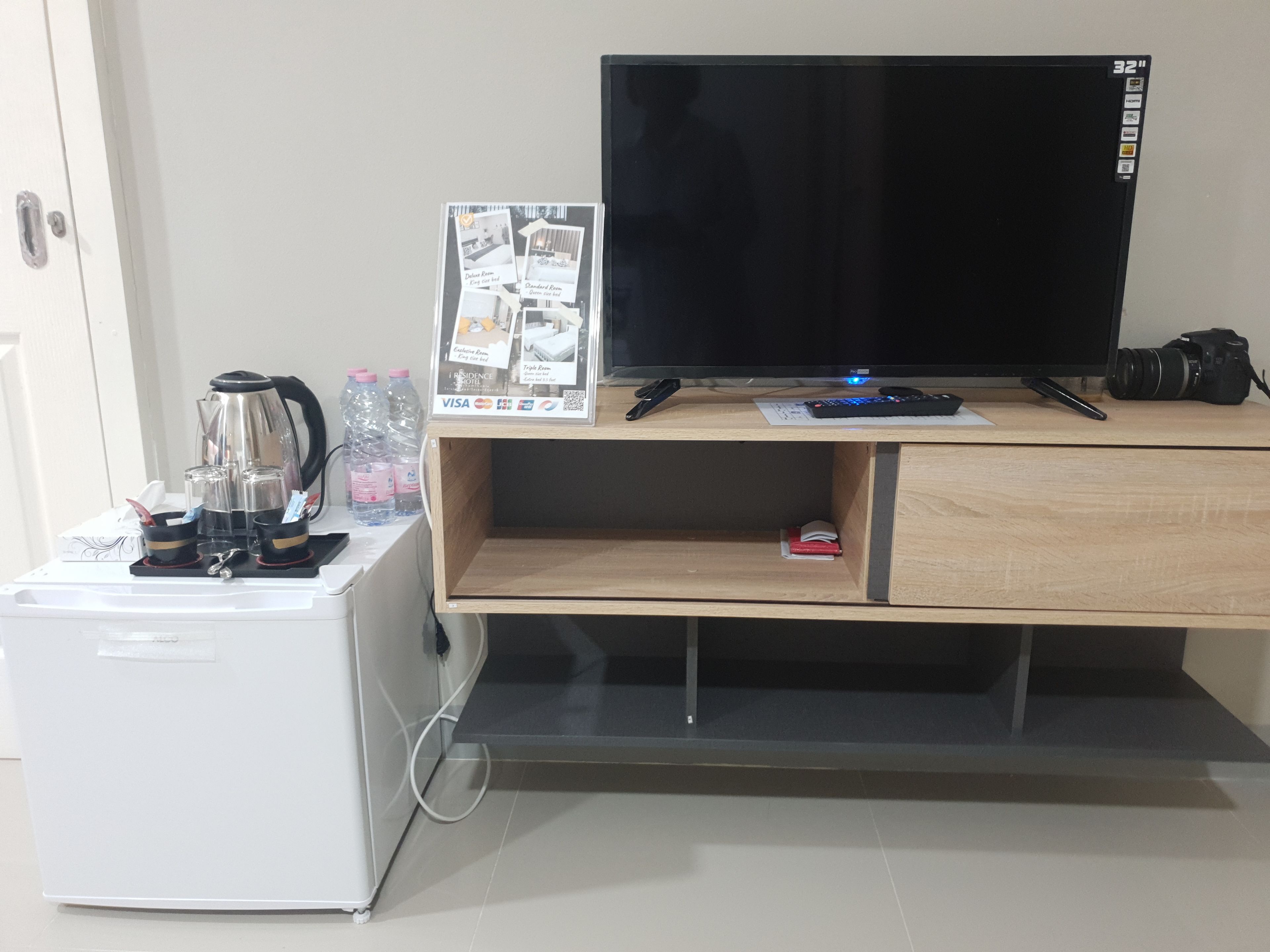 A compact kitchenette with a coffee maker, water bottles, and a small refrigerator. A flat-screen TV is mounted on a wooden cabinet above a desk with a camera and a remote control.