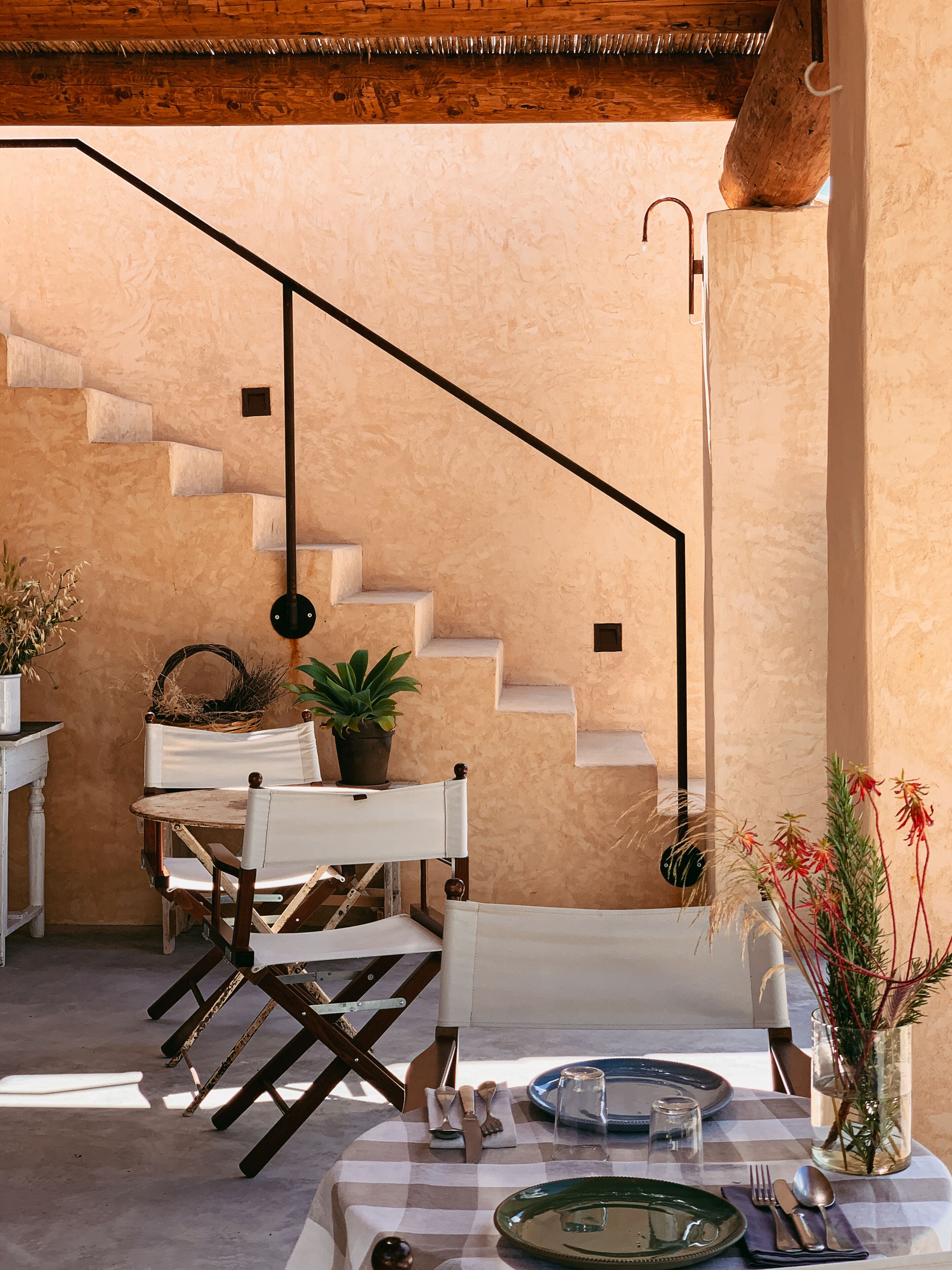 Un comedor con mantel a cuadros, una mesa puesta con cubiertos, y una escalera frente a una pared con textura.