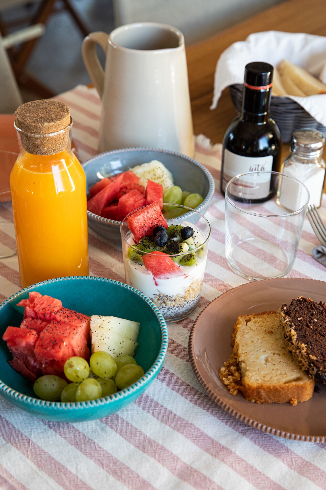 En morgenmadsborddækning med en kande, en skål med frugt, et glas juice og en skive brød på en ternet dug.