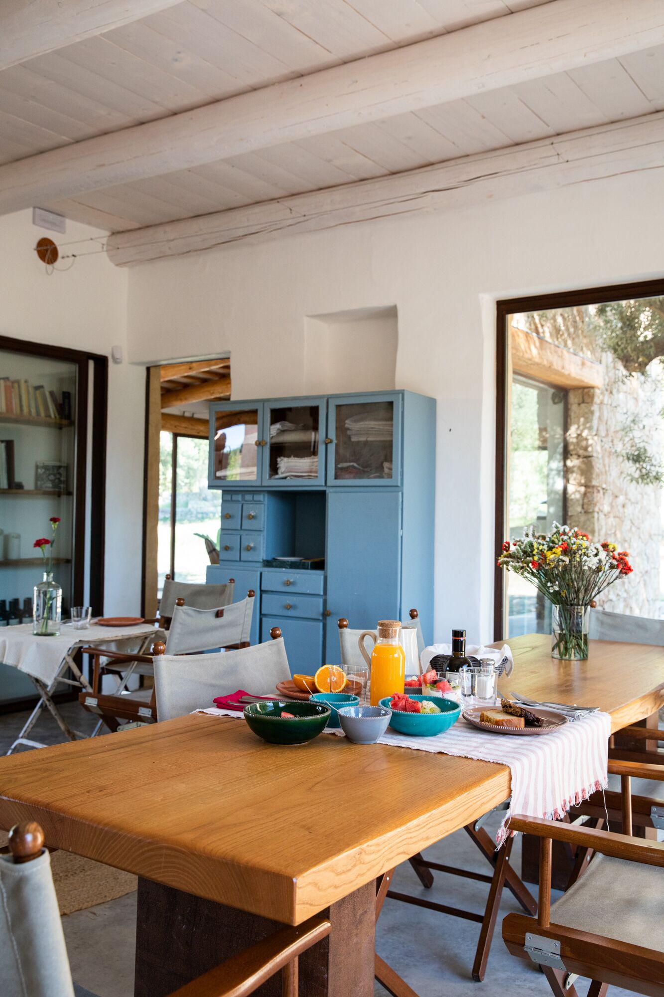 A dining area with a wooden table set for breakfast, a blue cabinet with glass doors, and a view of greenery outside.