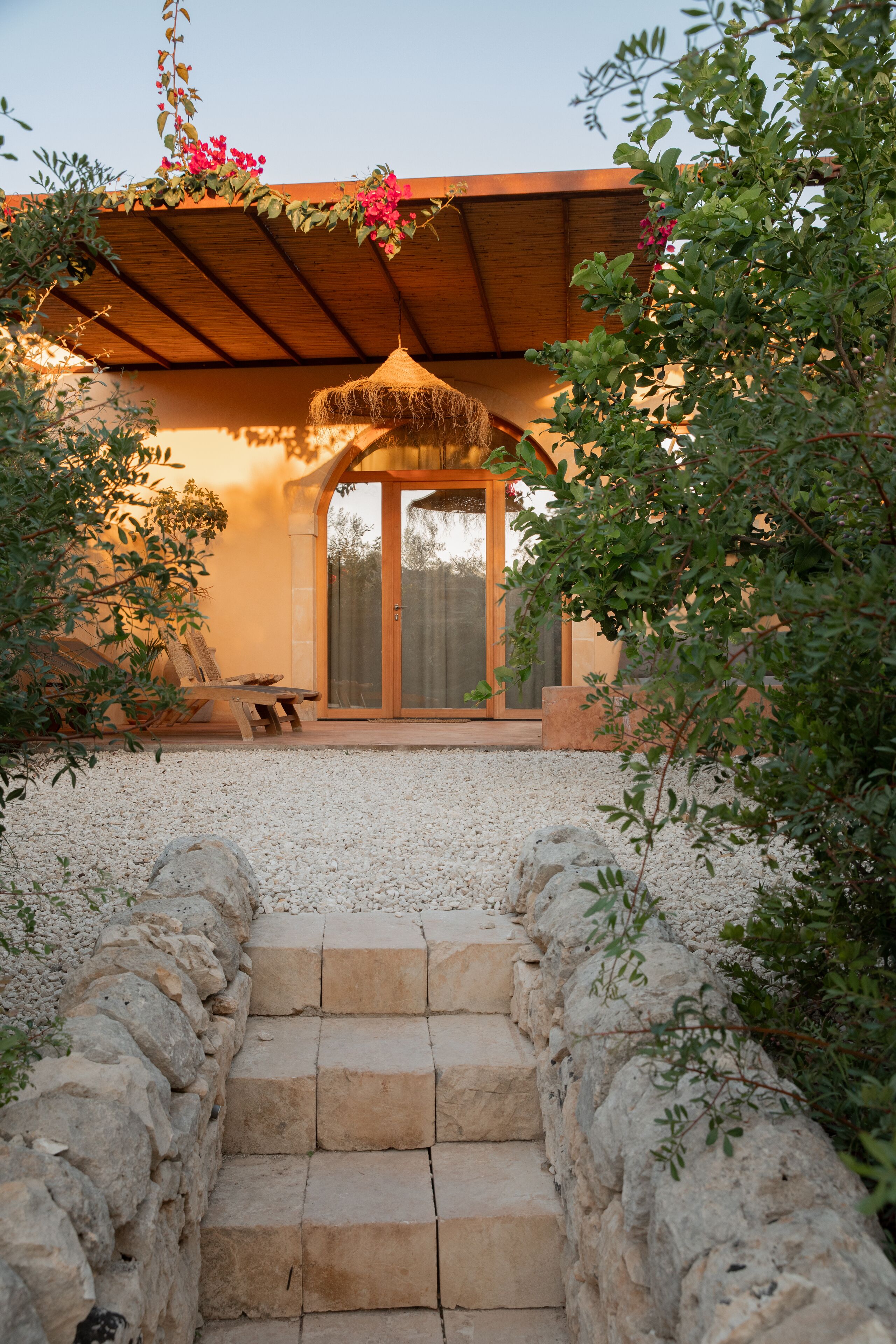 Una escalera de piedra que conduce a una puerta de madera con un panel de vidrio, rodeada de follaje y una estructura de techo de paja.