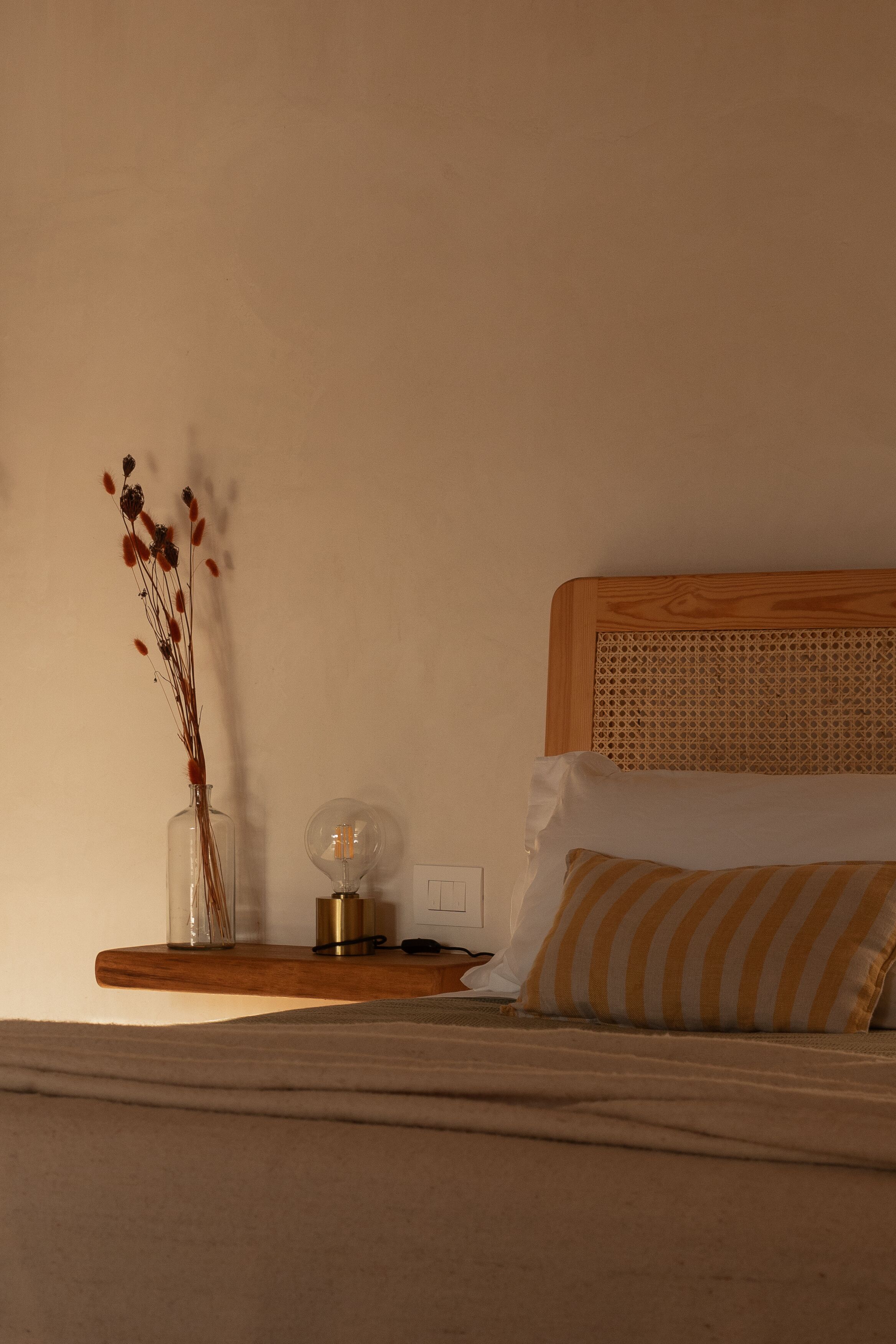 Un lit bien fait, avec un oreiller à rayures, une table de chevet surmontée d’un vase transparent et d’une bougie, et une tête de lit en bois.