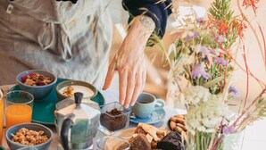 Uma mesa de pequeno-almoço com diversos alimentos, uma cafeteira e um vaso com flores.