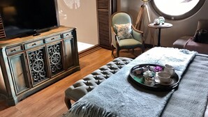 A cozy room with a television on a wooden cabinet, a round window, a chair with a cushion, and a tray with cups and a teapot on a bed. - Regnum Golf Country 3 bedroom 4 (Bodrum)