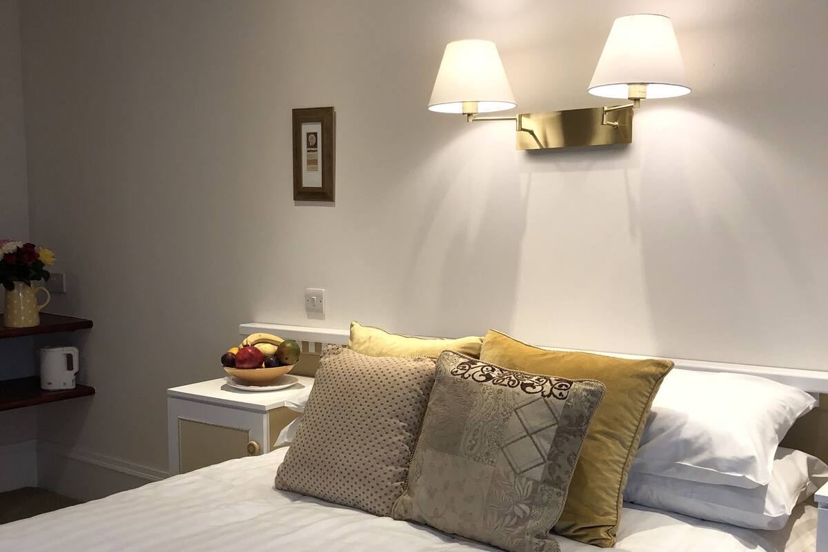 Cama bem arrumada com roupa de cama branca, duas luminárias na parede, uma mesinha de cabeceira com uma fruteira e um quadro na parede.