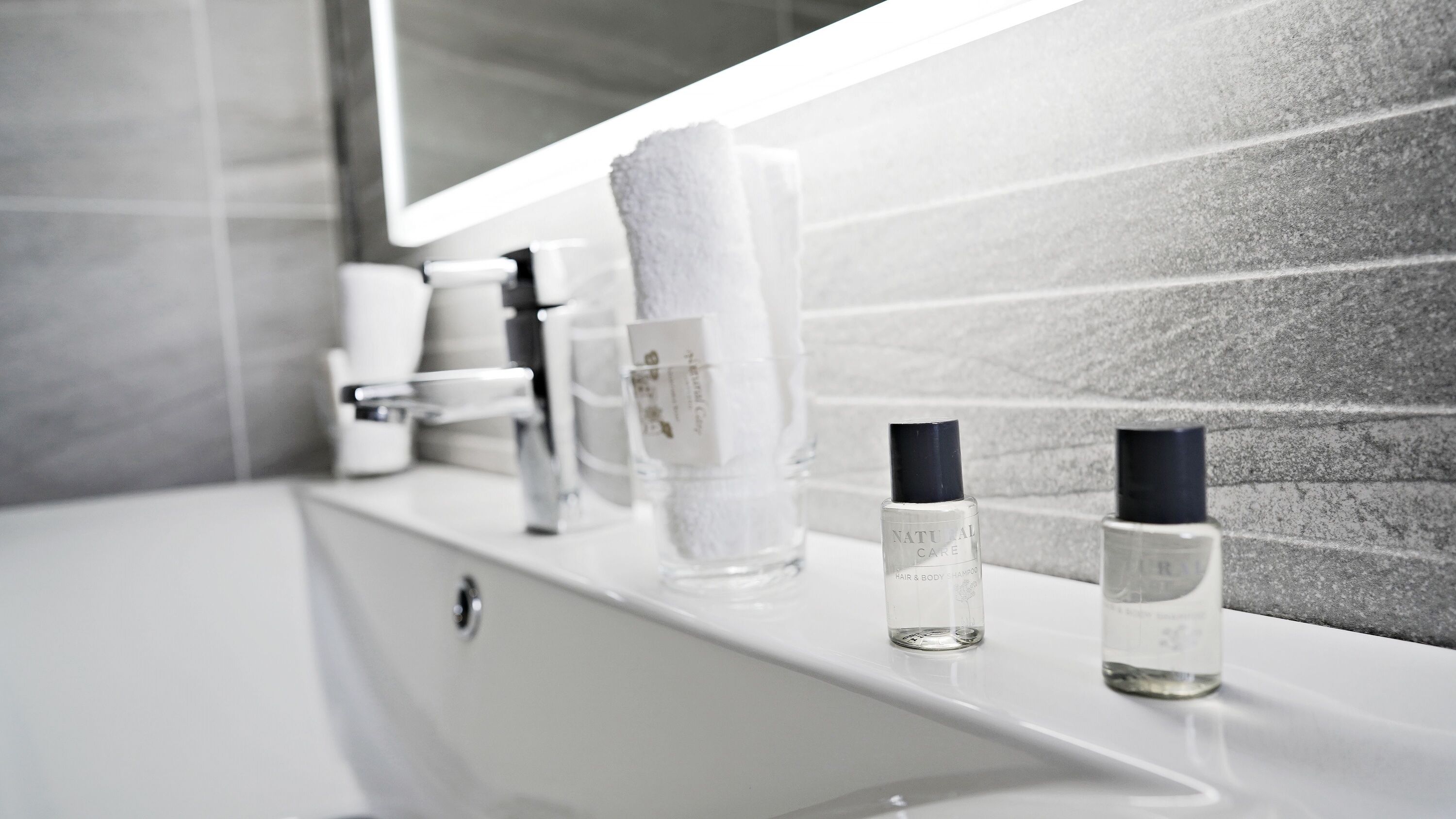 A bathroom with a white sink, a mirror, and toiletries including a bottle of shampoo and a glass of water.