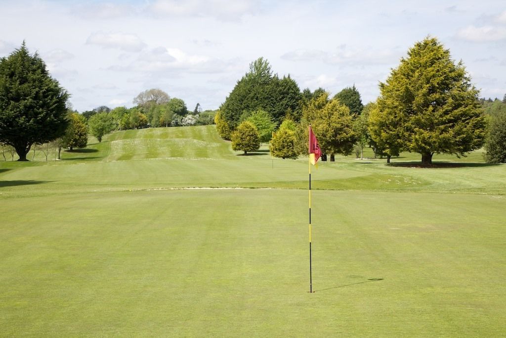 Un campo de golf bien cuidado, con un marcador de posición en primer plano, rodeado de árboles frondosos y colinas onduladas.
