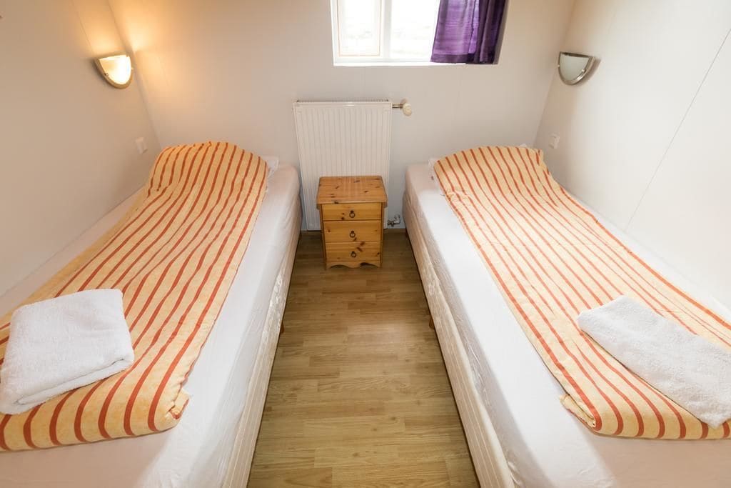 A small, clean room with two single beds, each with a striped bedspread and a white towel. There is a wooden chest of drawers in the center and a window with curtains on the wall.