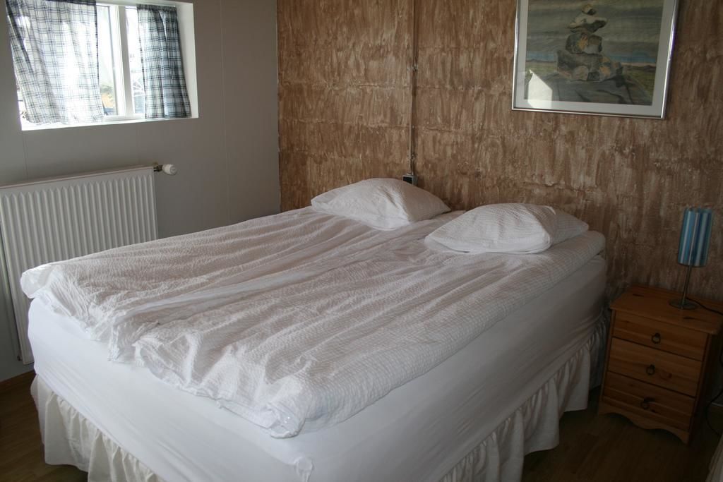A bedroom with a large bed, a wooden headboard, a window with curtains, a radiator, a bedside table, and a drawer.