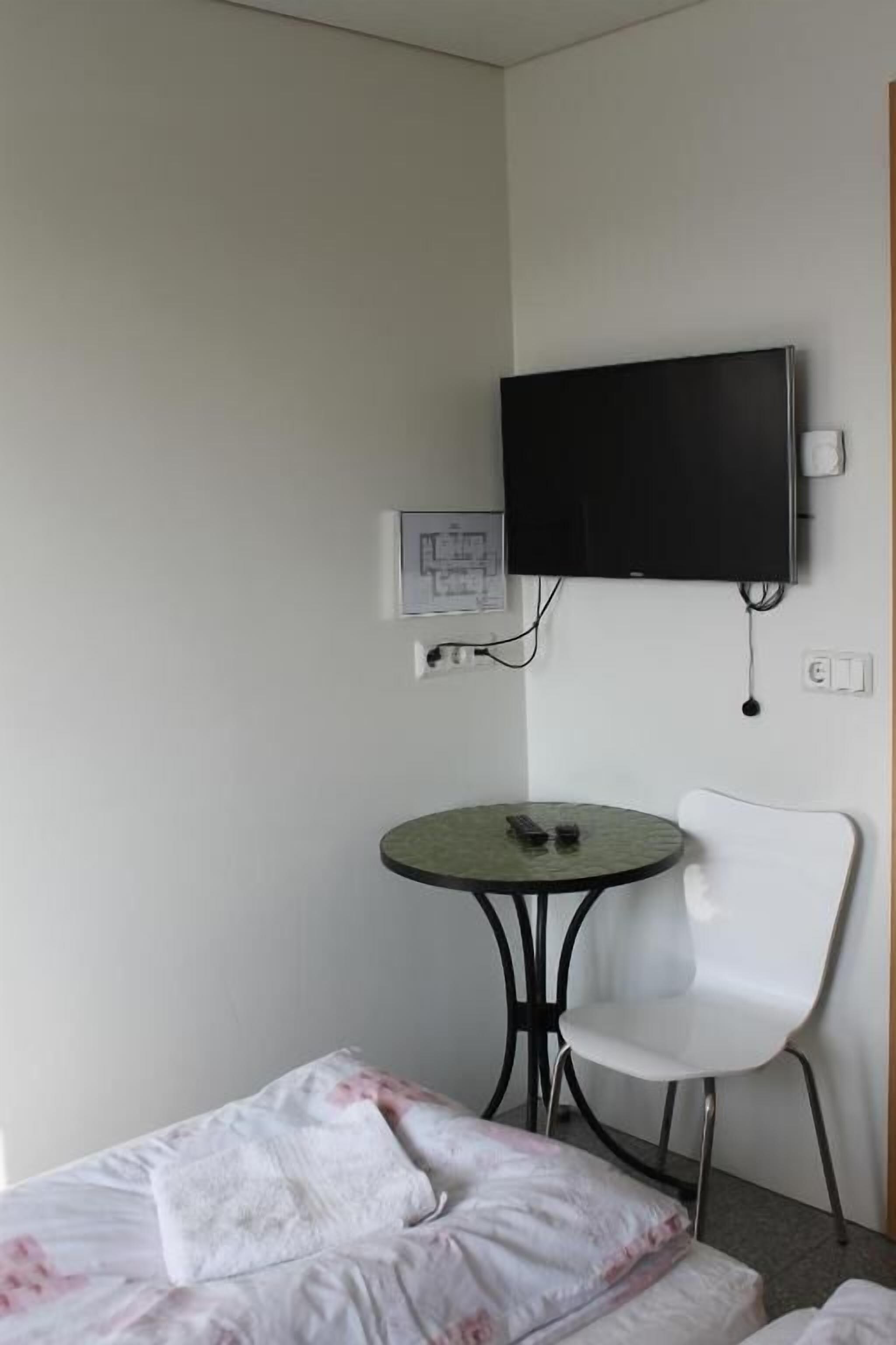 Una habitación pequeña con una cama, una silla blanca, una mesa redonda y un televisor montado en la pared.