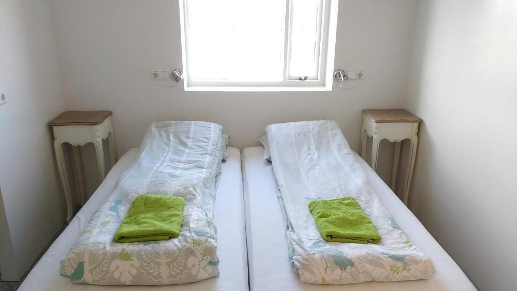 Une chambre avec deux lits simples, chacun agrémenté d’une serviette verte, une fenêtre au-dessus et des tables de chevet.