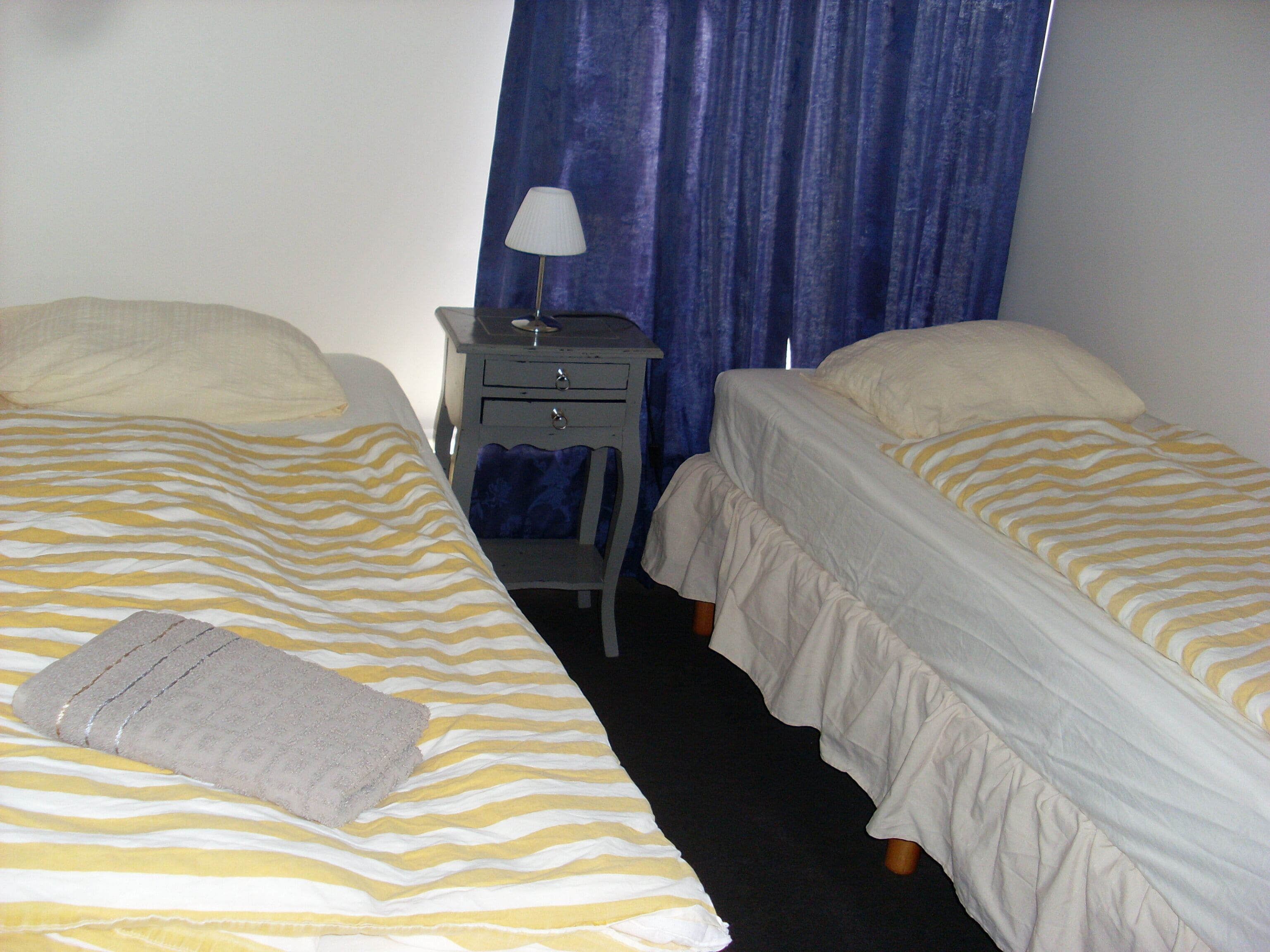 Habitación de hotel con dos camas, una mesita de noche, una lámpara y una ventana con cortinas azules.