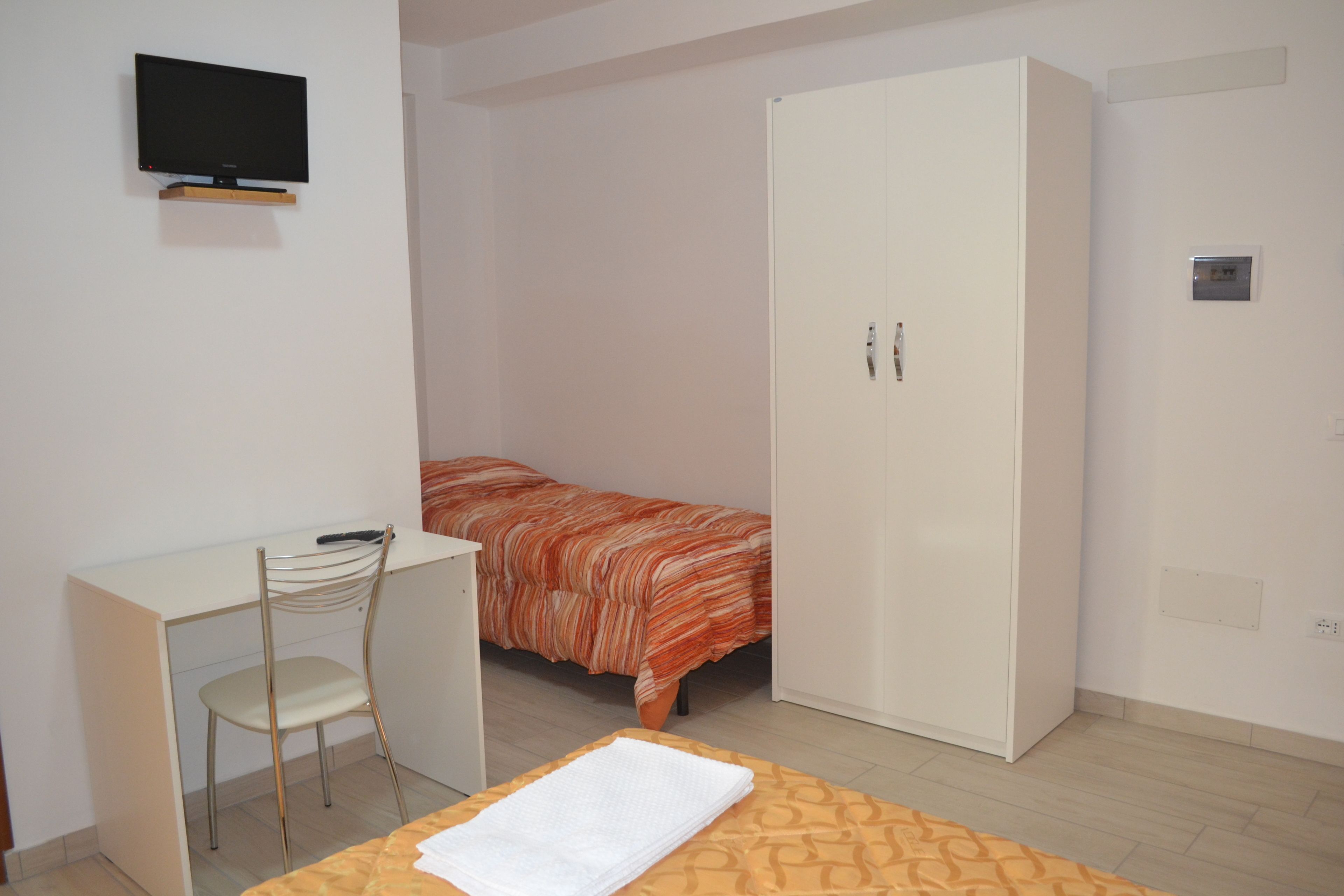 A bedroom with a bed, a desk, a chair, a wardrobe, and a television on the wall.