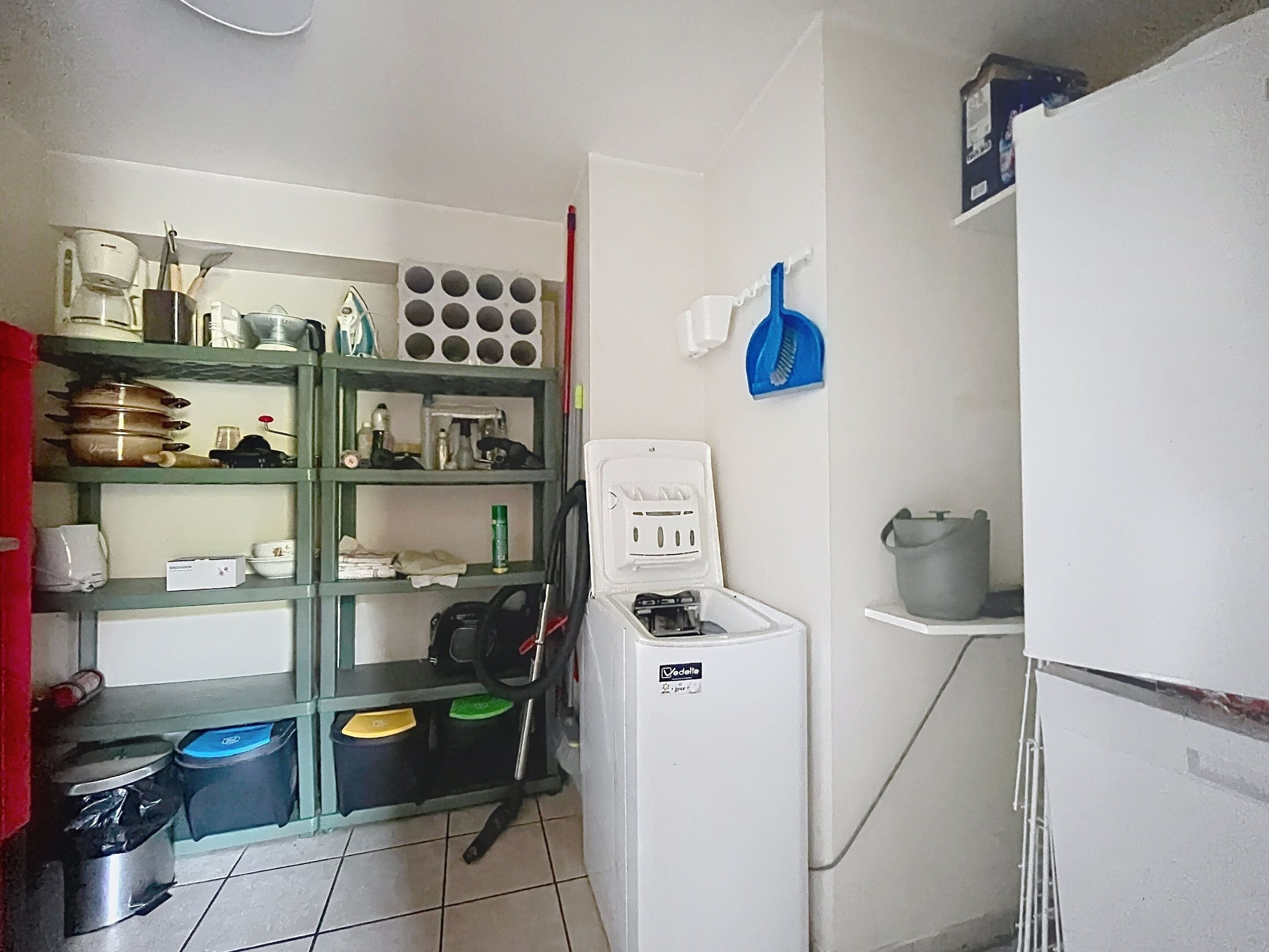 A kitchen with shelves, a refrigerator, and a trash bin.