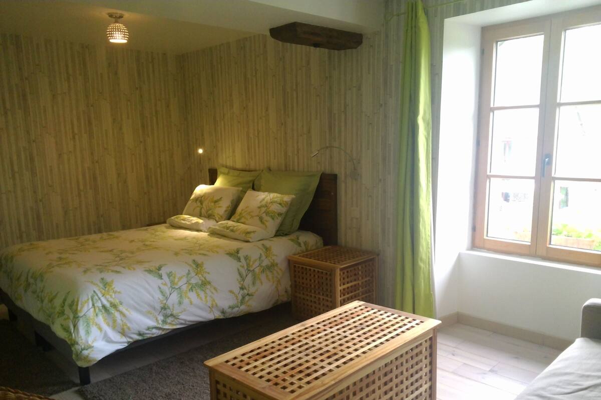 A bedroom with a bed, a wooden headboard, a nightstand, a window with curtains, and a wooden floor.