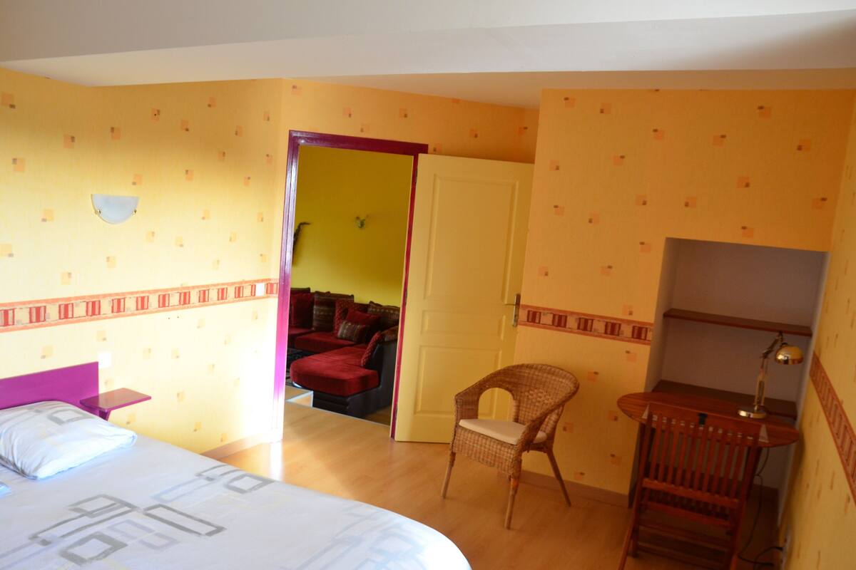 A hotel room with a bed, a chair, a desk with a lamp, and a door leading to another room.