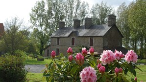Un bâtiment en pierre, doté d’un toit symétrique et de plusieurs cheminées, entouré d’une végétation luxuriante et de plantes en fleurs.