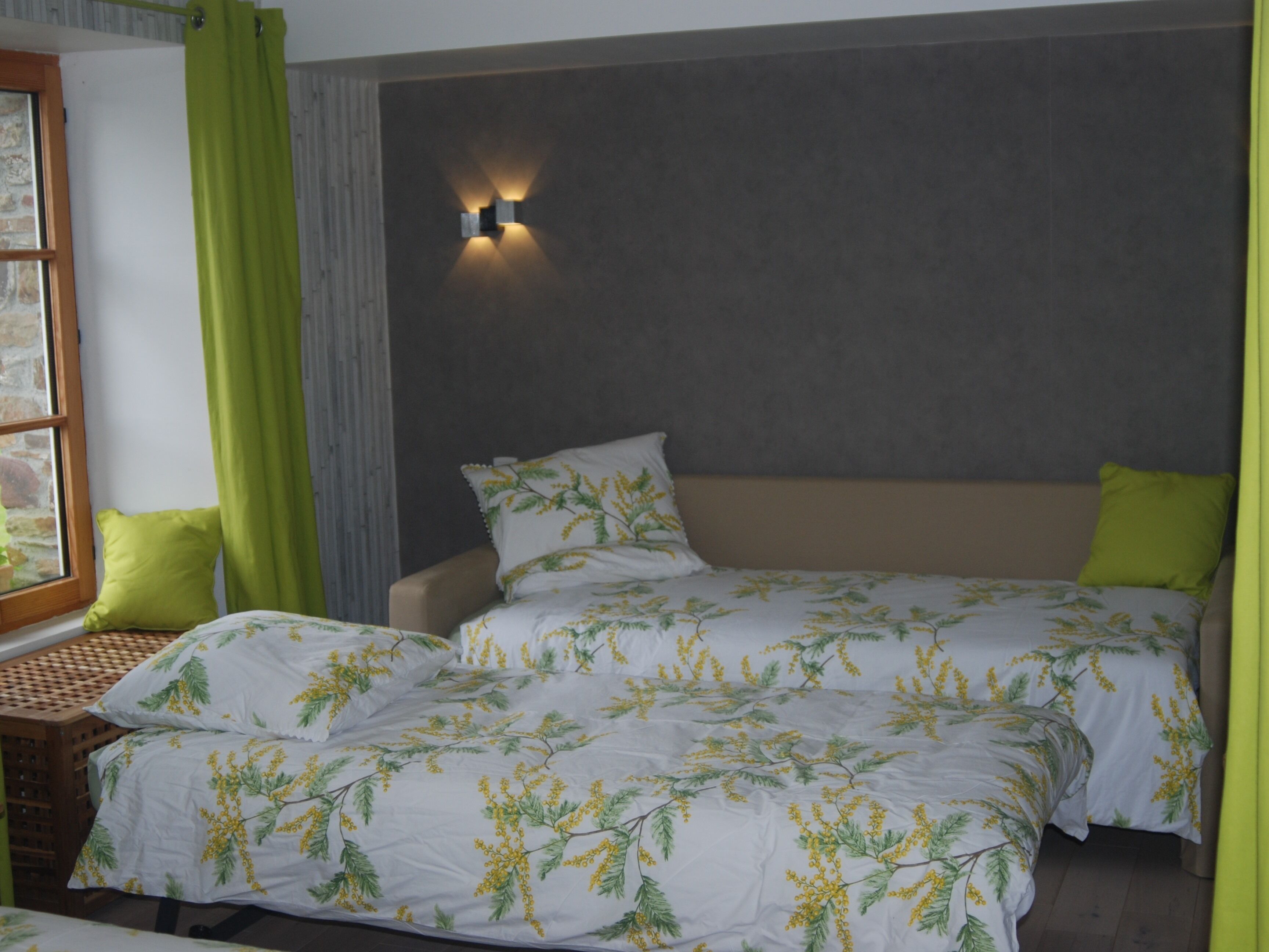 A bedroom with two beds, a nightstand, a window with curtains, and a wall-mounted light fixture.