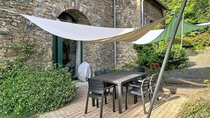 Un bâtiment en pierre avec une terrasse en briques, des sièges extérieurs et de la verdure.