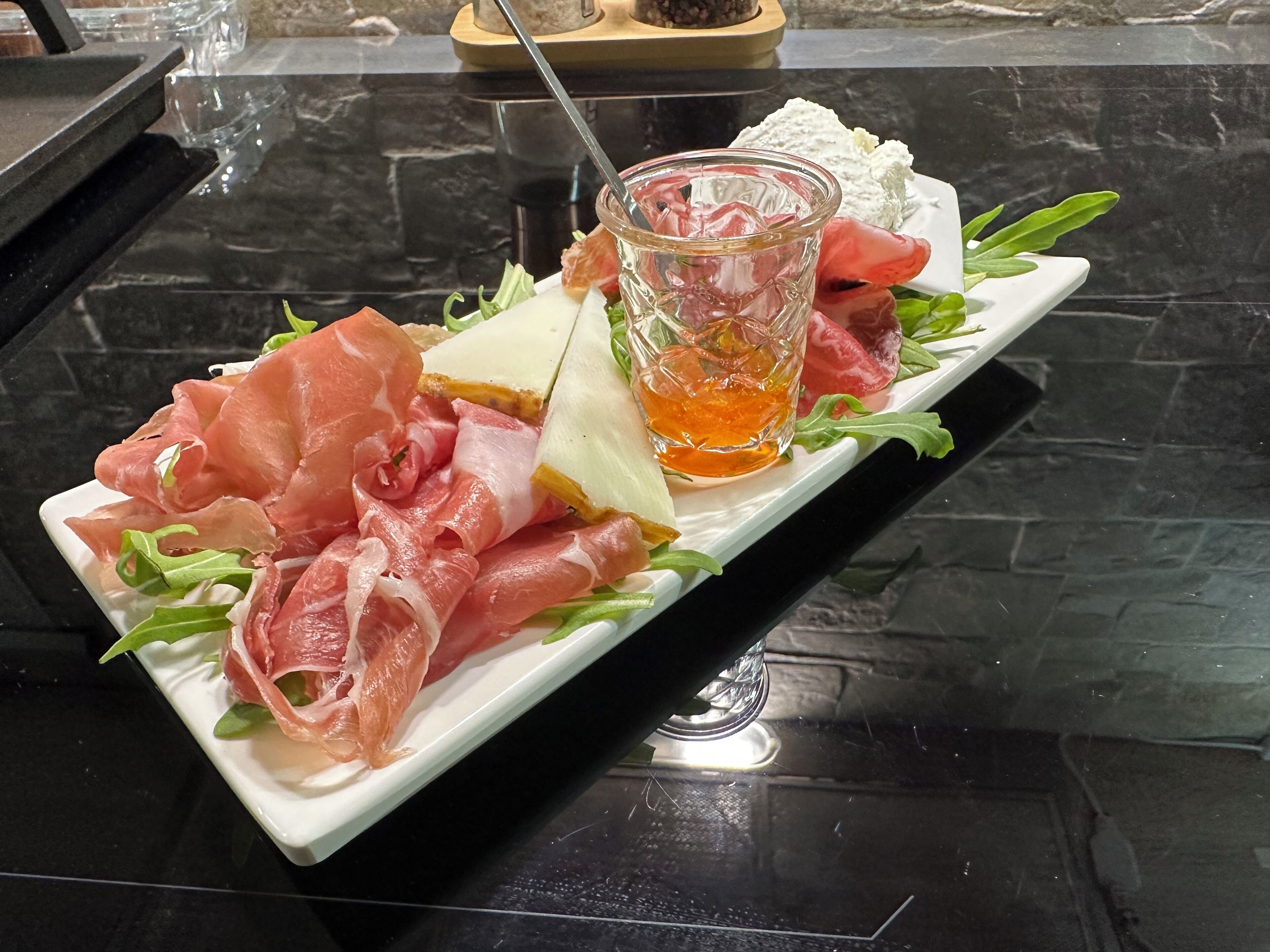 A rectangular white plate with various food items including prosciutto, cheese, and a glass of orange liquid, likely a drink, on a black countertop.