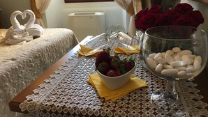 A neatly made bed with a heart-shaped towel swan, a table with lace tablecloth, a bowl of strawberries, a glass of white stones, and red roses. - B&B Sei Stelle (Sulmona)