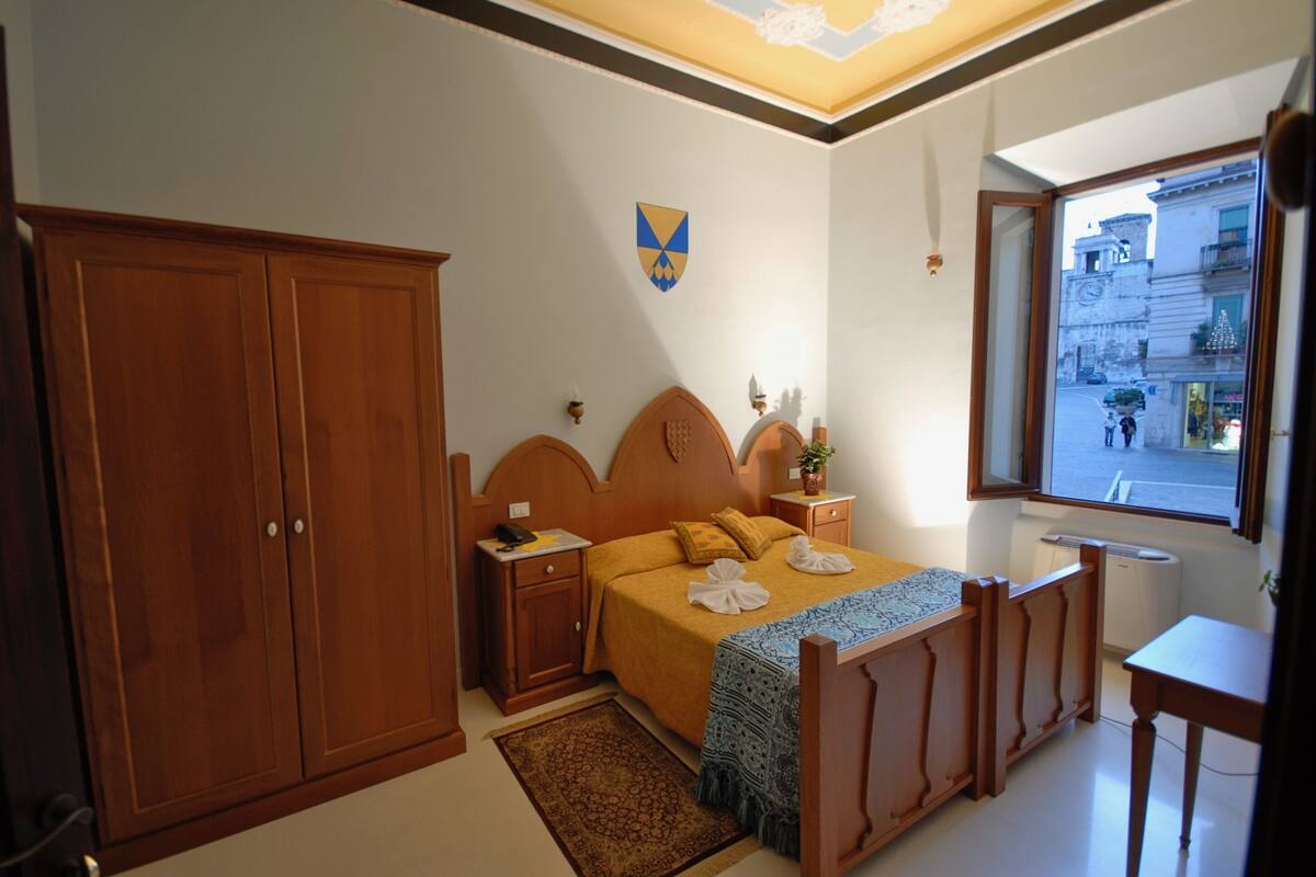 A bedroom with a bed, wooden wardrobe, bedside tables, a window with a view of the street, and a ceiling with a decorative pattern.