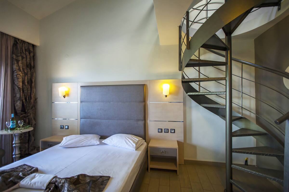 A hotel room with a bed, bedside tables, a lamp, a small table with a bottle and glasses, and a spiral staircase.