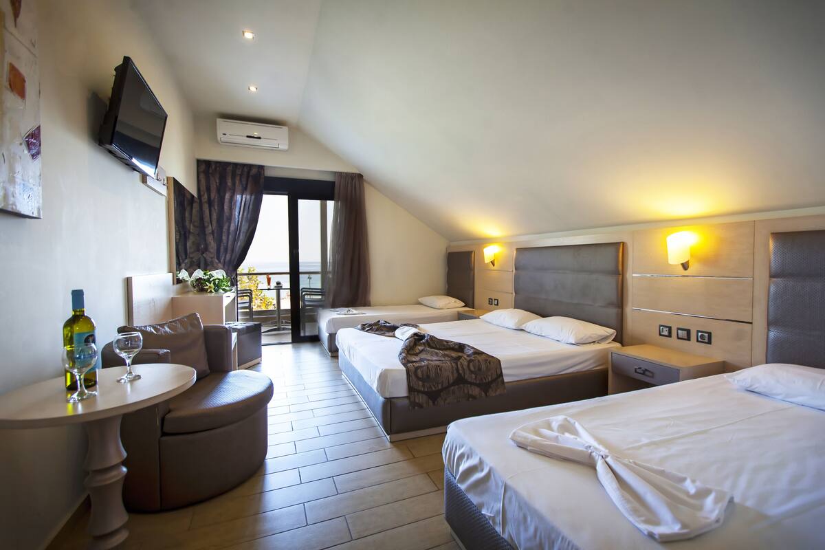 A hotel room with two beds, a sofa, a small table with a bottle and glass, and a TV mounted on the wall.