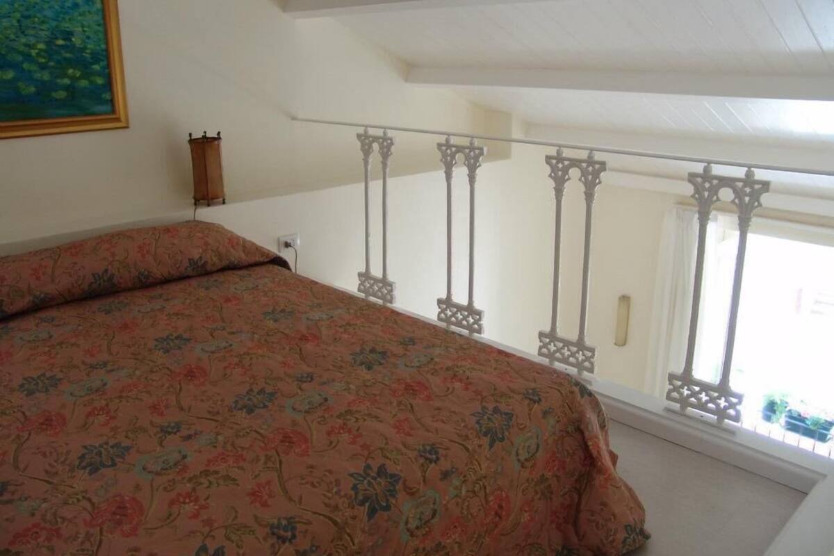 A bedroom with a bed, a painting on the wall, and a balcony with a railing.