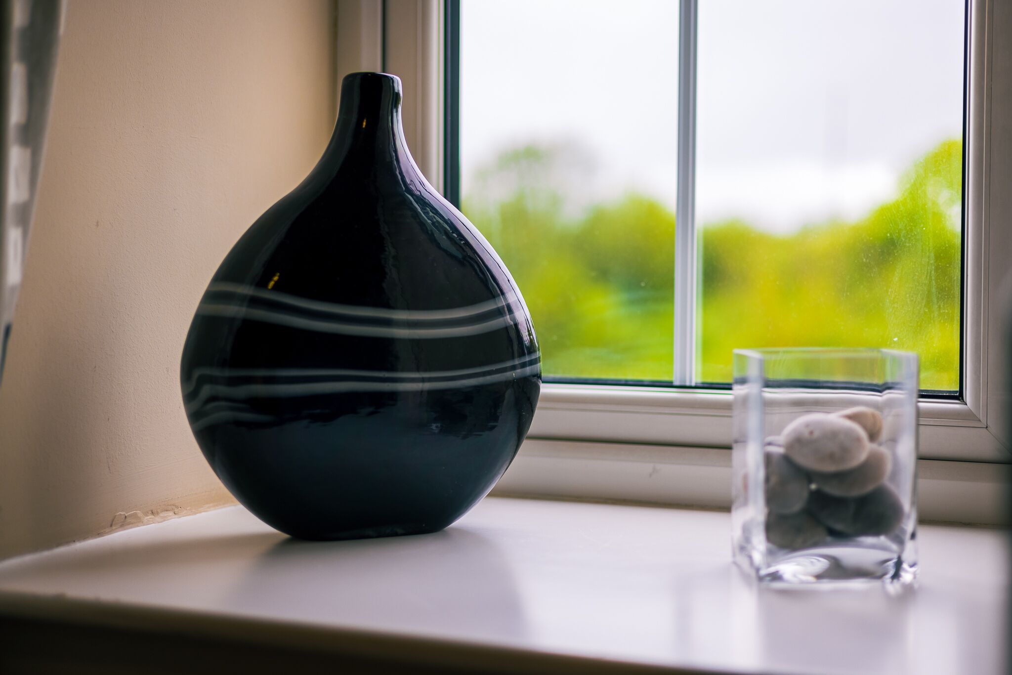 A black vase with white stripes on a windowsill next to a clear glass containing stones.
