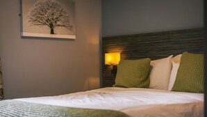 A neatly made bed with white linens and a green throw blanket, a wall-mounted lamp with a yellow shade, and a framed picture of a tree. - The Old Mill (Durham)