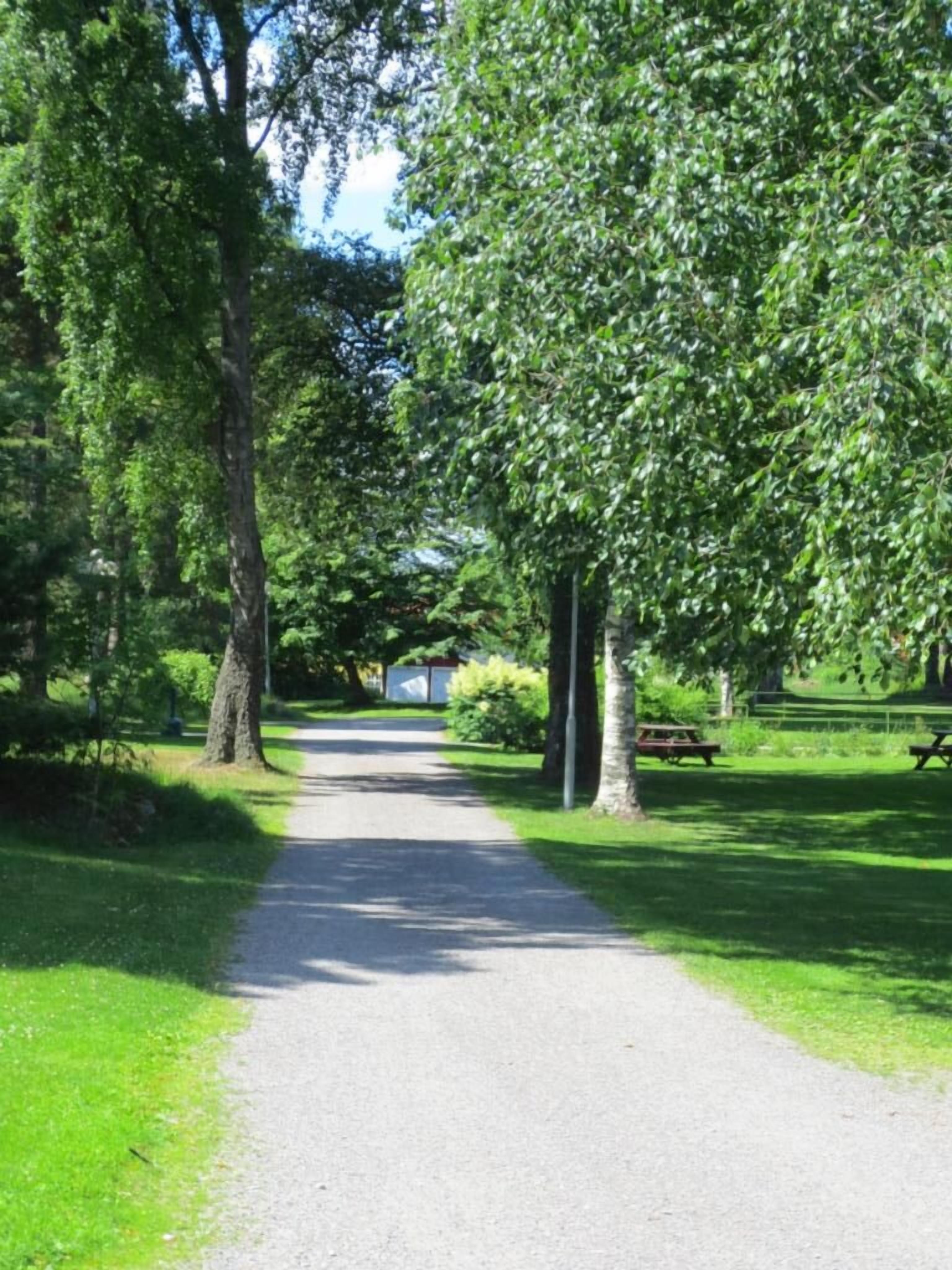 Asfaltoitu kävelytie, jonka molemmin puolin on puita ja nurmikkoa, johtaa rakennukseen, jonka yhteydessä on piknikpöytä.