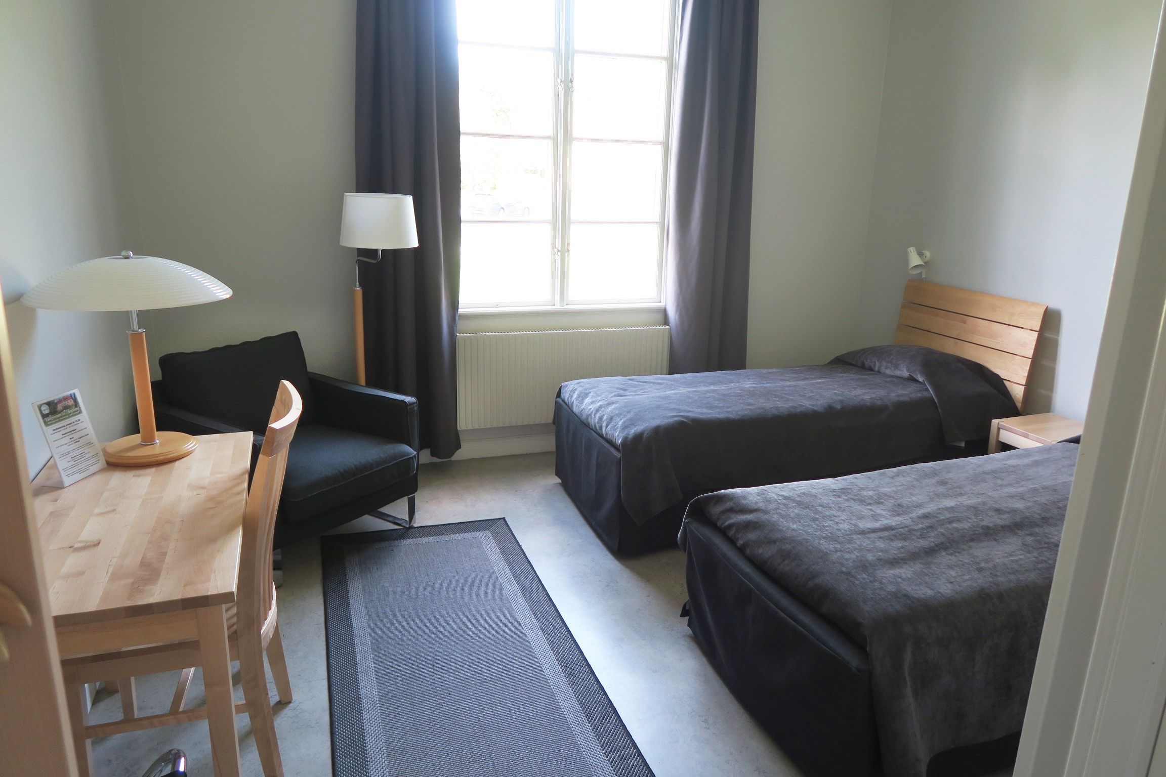 A hotel room with two beds, a desk, a chair, a lamp, and a window with curtains.
