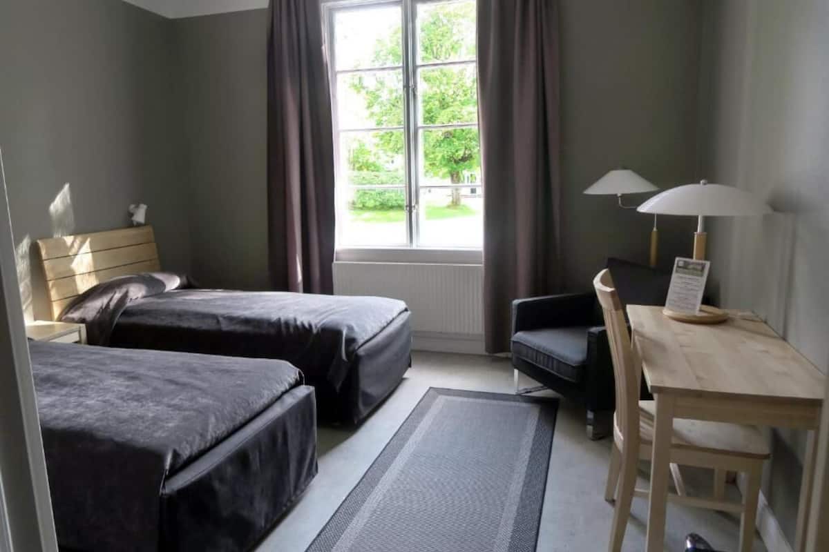 A room with two beds, a desk, a chair, a window with curtains, and a lamp.
