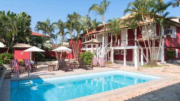 A swimming pool with lounge chairs and umbrellas, surrounded by palm trees and a red building with a tiled roof. - Pousada Tartaruga (Búzios)