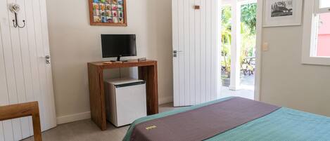 A bedroom with a bed, a small desk with a computer, a refrigerator, a wooden chair, a door leading outside, and a wall with a framed picture.