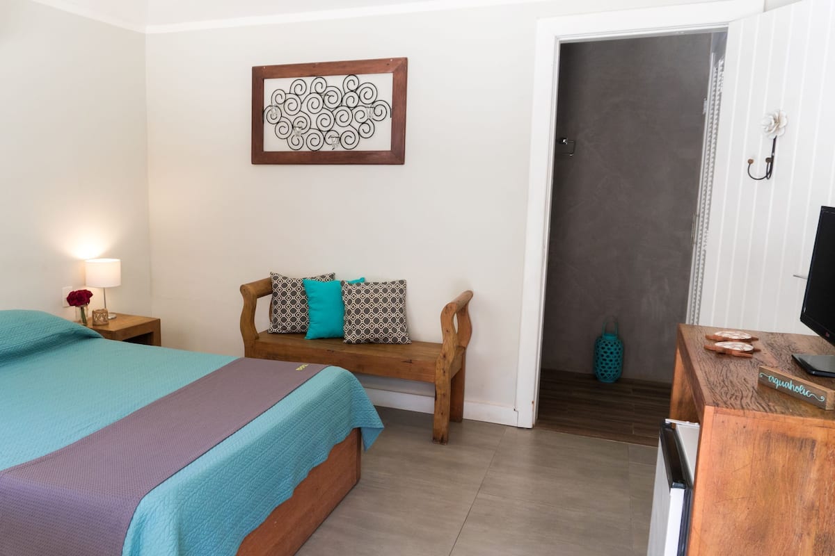 A bedroom with a bed, a wooden bench with cushions, a wall-mounted artwork, and a desk with a computer and a sign.