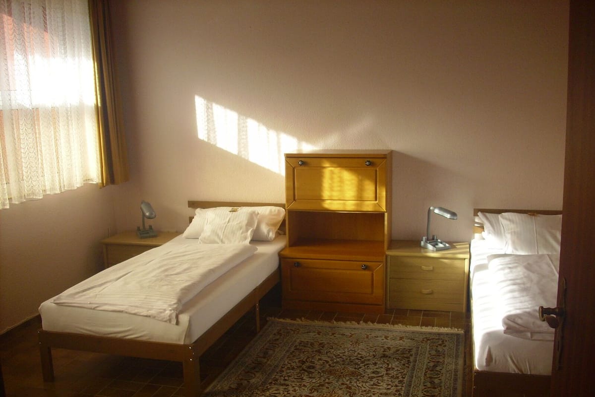 A small, neatly arranged bedroom with a single bed, a nightstand, a dresser, and a desk with a lamp.