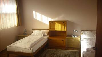 Ein kleines, ordentlich eingerichtetes Schlafzimmer mit einem Einzelbett, einem Nachttisch, einer Kommode und einem Schreibtisch mit einer Lampe.